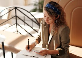 Woman in blazer writing notes while using smartphone in sunlit cafe.